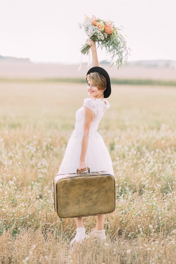 The side view of the smiling bride carrying the vintage suitcase, keeping up the colourful bouquet and looking at the