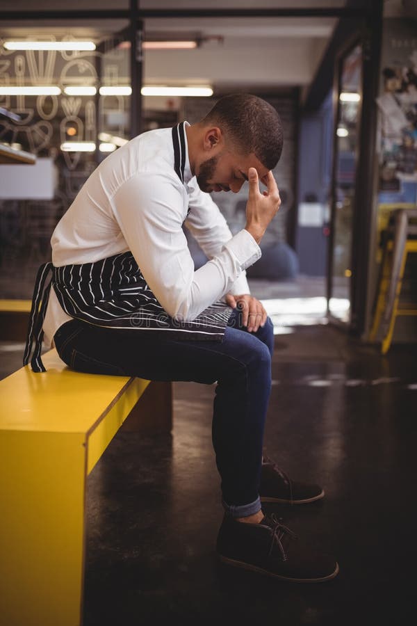 Side view of upset young waiter sitting on yellow bench with headache
