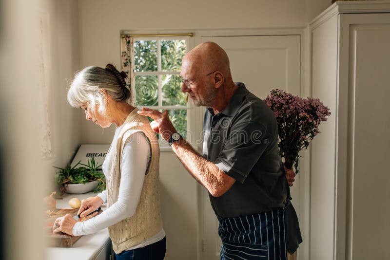 Side view of a woman working in kitchen with her husband standing behind with a bouquet of flowers. Senior man hiding a bunch of stock photography Side view of a woman working in kitchen with her husband standing behind with a bouquet of flowers. Senior man hiding a bunch of stock photography
