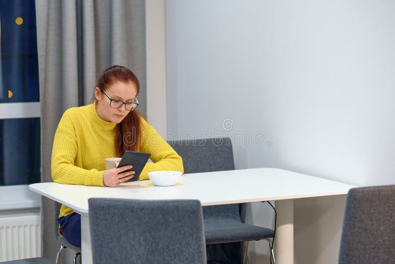 Side View Of The Young Caucasian Woman In Yellow Knitted Sweater Seats At The Table In Modern Light Interior And Reading An