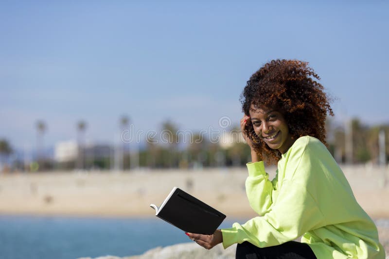 Side view of young curly afro woman sitting on a breakwater holding a book while smiling and looking away outdoors royalty free stock photo Side view of young curly afro woman sitting on a breakwater holding a book while smiling and looking away outdoors royalty free stock photo