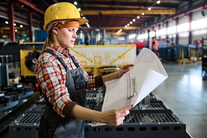 Side view of young female engineer in workwear holding unrolled blueprint royalty free stock image