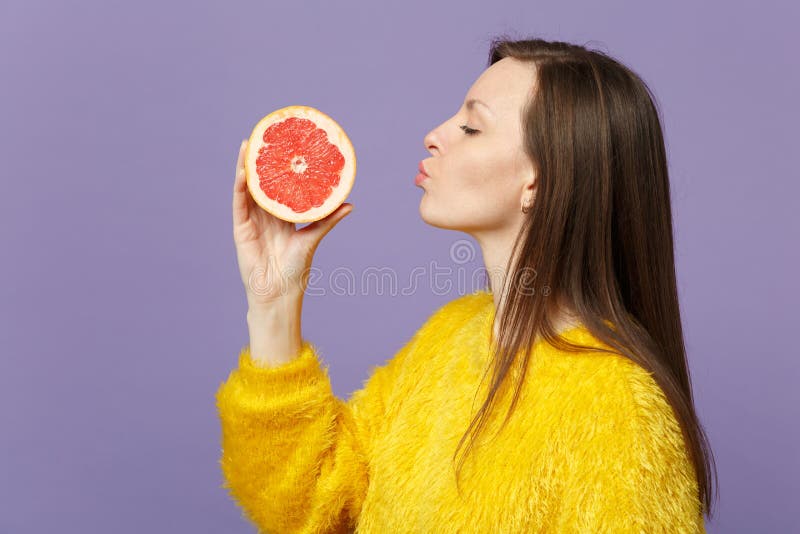 Side view of young woman in fur sweater hold sending air kiss to half of fresh ripe grapefruit isolated on violet pastel stock photography