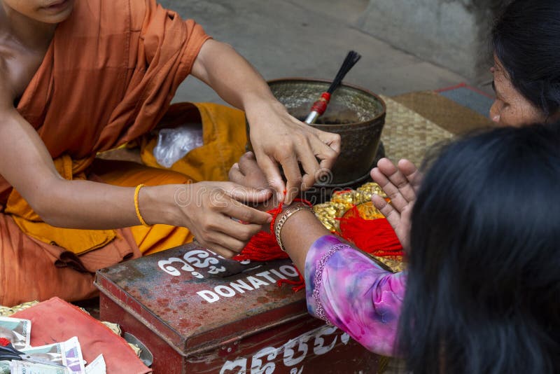 Siem Reap, Cambodia - 25 March 2018: Buddhist monks in orange clothes in Angkor Wat temple. Buddhist ritual of reading mantra and giving red string. Buddhist bracelet from red thread. Angkor Wat scene. Siem Reap, Cambodia - 25 March 2018: Buddhist monks in orange clothes in Angkor Wat temple. Buddhist ritual of reading mantra and giving red string. Buddhist bracelet from red thread. Angkor Wat scene