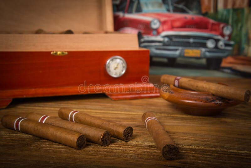 Cuban cigars and humidor with ashtray on rustic wooden table with Cuban painting of american old car in background. Cuban cigars and humidor with ashtray on rustic wooden table with Cuban painting of american old car in background