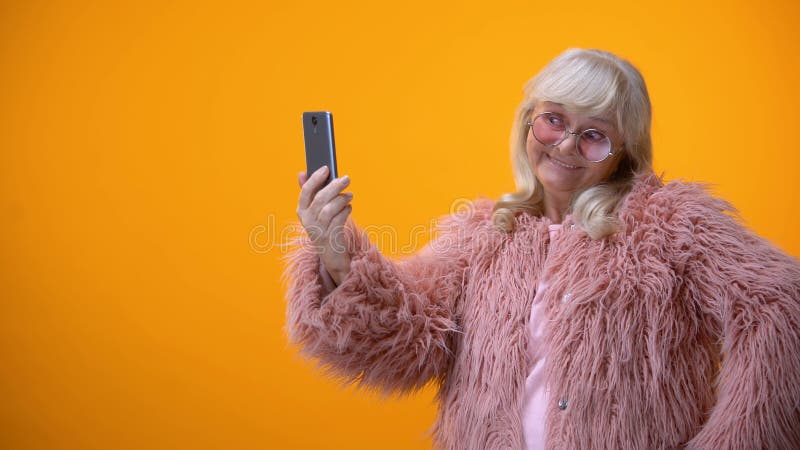 Signora senior allegra in cappotto rosa divertente che prende selfie sullo smartphone, divertendosi