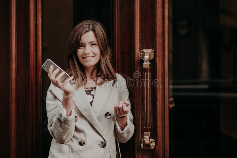 Signora splendida elegante in impermeabile, le tenute il telefono cellulare, gli aspettare la chiamata, porte vicine all'aperto d immagini stock Signora splendida elegante in impermeabile, le tenute il telefono cellulare, gli aspettare la chiamata, porte vicine all'aperto d immagini stock