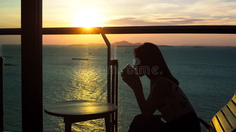 Silhouet van een mooie Aziatische vrouw die een kop van koffie met warme middagzonsondergang drinken bij de oceaan op de achtergr royalty-vrije stock foto Silhouet van een mooie Aziatische vrouw die een kop van koffie met warme middagzonsondergang drinken bij de oceaan op de achtergr royalty-vrije stock foto