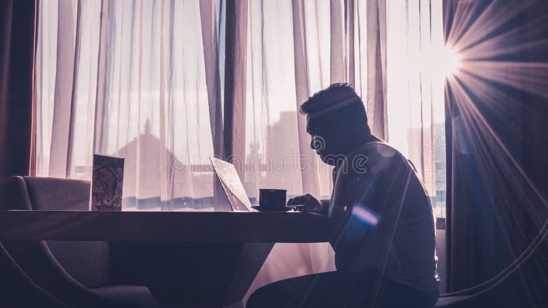 The silhouette of a man at work using a laptop in a modern room with a window background illuminated by slightly dazzling natural sun glare. The silhouette of a man at work using a laptop in a modern room with a window background illuminated by slightly dazzling natural sun glare