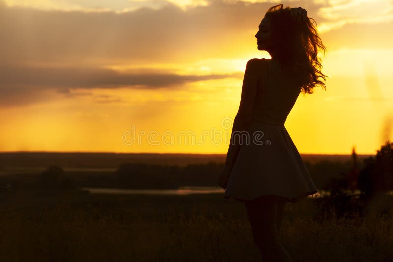 Silhouette of a beautiful girl in a dress at sunset in a field, figure of young woman enjoying nature, concept of leisure