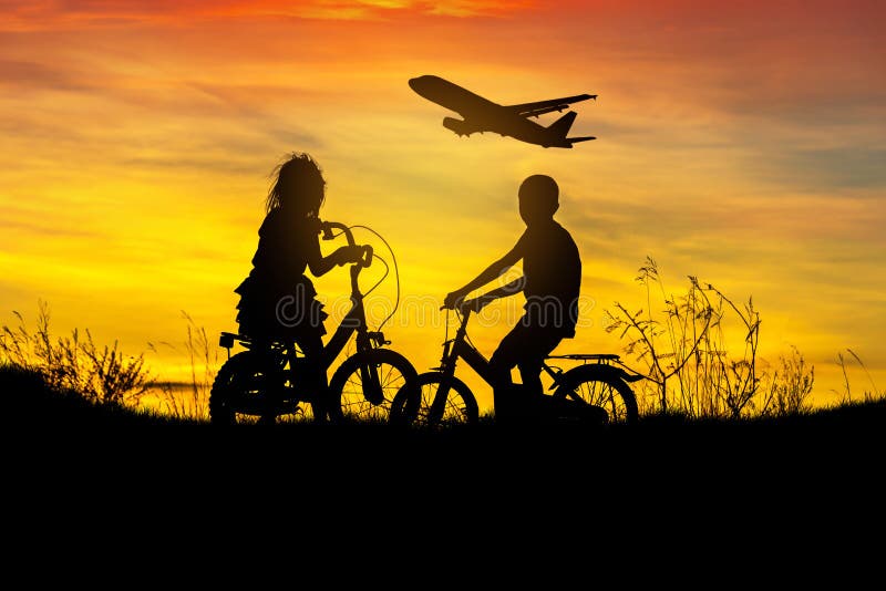 Silhouette little boy and little girl riding bike look to airplane flying on sunset