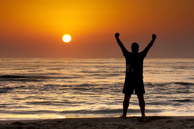 Silhouette di giovane uomo in aumento braccia sulla spiaggia al sorgere del sole.
