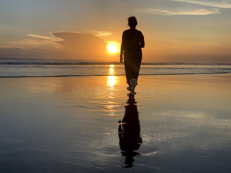 Silhouette of a woman walking alone with bare feet in the beach at sunset.