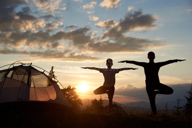 Silhouette of woman and boy standing in yoga pose at sunset near the camping under beautiful sky