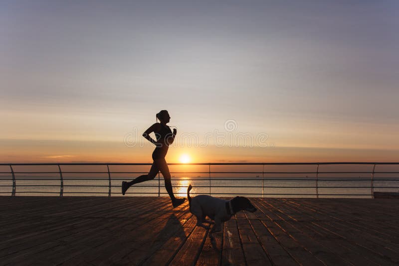 Silhouette of a young beautiful athletic girl with long blond hair in headphones that runs at dawn over the sea with her dog stock photos Silhouette of a young beautiful athletic girl with long blond hair in headphones that runs at dawn over the sea with her dog stock photos