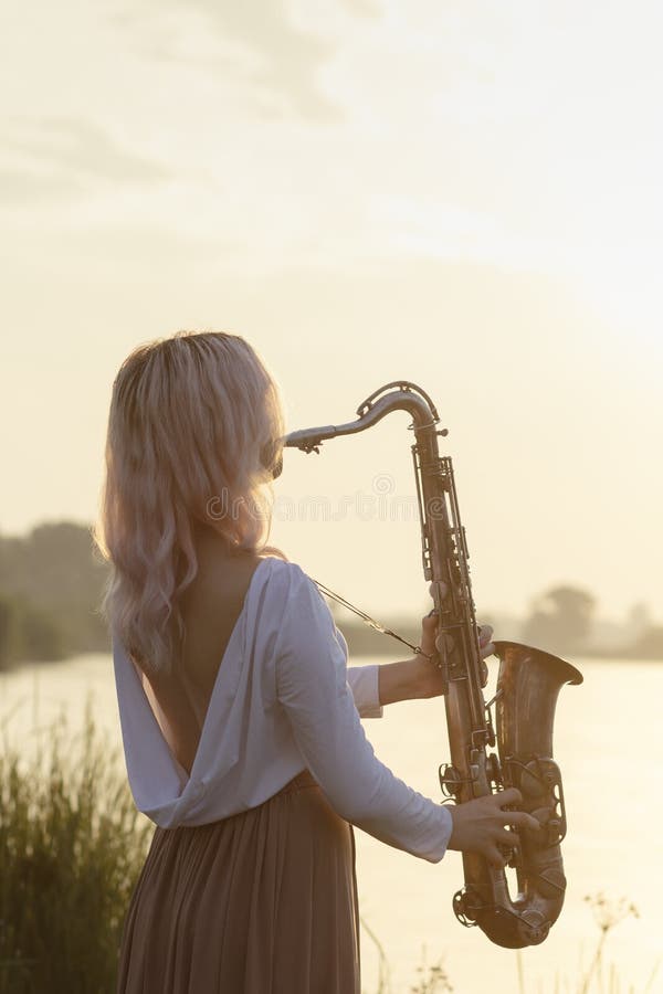 Silhouette of a young beautiful girl playing the saxophone at sunrise by the river, a woman in a long dress on the nature at