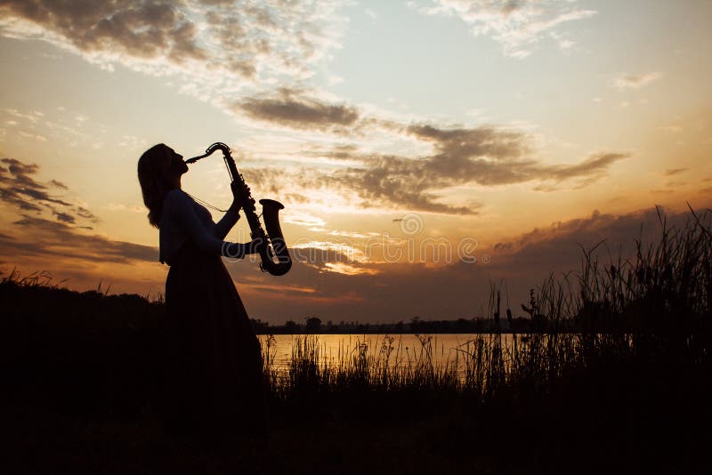 Silhouette of a young beautiful girl playing the saxophone at sunrise by the river, a woman in a long dress on the nature at