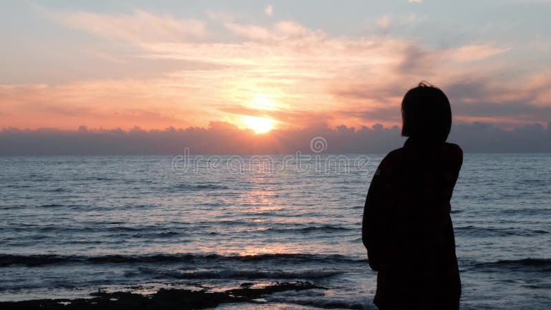 Silhouette of young girl looking at sunset. Attractive girl in casual wear standing on rocky shore and looking at ocean. Silhouett