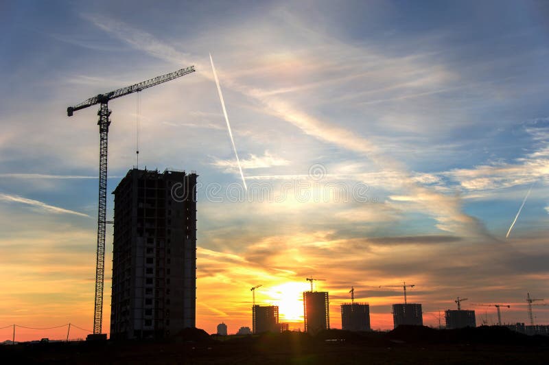 Silhouettes of tower cranes constructing a new residential building at a construction site against sunset background.