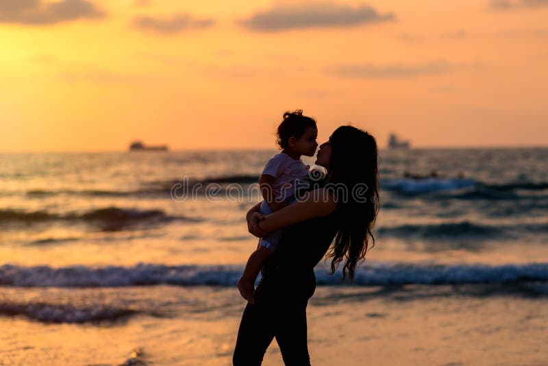 Silhouettes young mother with daughter playing and kissing on the beach at sunset evening sky background. Happy family. royalty free stock photo Silhouettes young mother with daughter playing and kissing on the beach at sunset evening sky background. Happy family. royalty free stock photo
