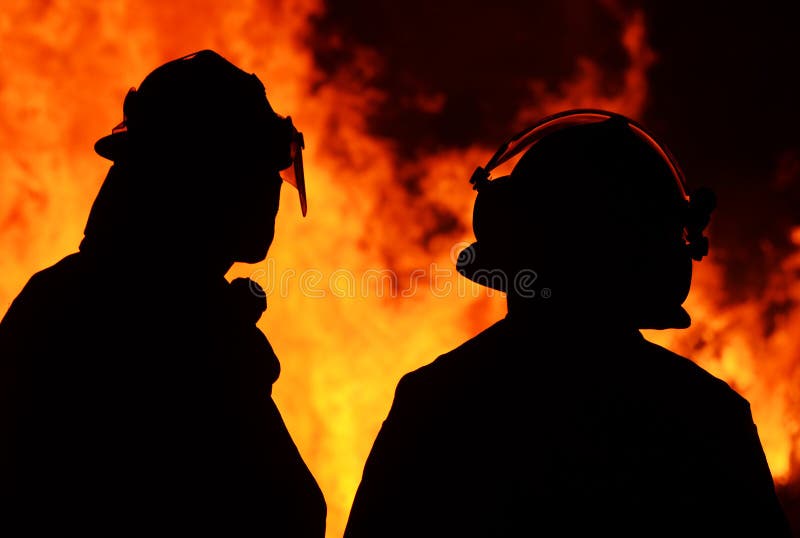 Silhouettes of a pair of brave firemen dressed in their uniform and safety gear with the raging flames of a huge bushfire in the background. Photo taken in the rural countryside of Queensland, Australia. Silhouettes of a pair of brave firemen dressed in their uniform and safety gear with the raging flames of a huge bushfire in the background. Photo taken in the rural countryside of Queensland, Australia.