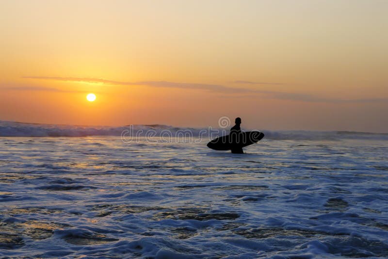 Silhouette of unknown anonymous surfer man holding surf board after surfing on sunset with amazing beautiful sunlight with orange sky and silver sea water in Summer sport and holiday vacation concept. Silhouette of unknown anonymous surfer man holding surf board after surfing on sunset with amazing beautiful sunlight with orange sky and silver sea water in Summer sport and holiday vacation concept