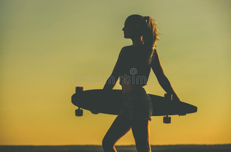 Silhueta de uma menina bonita, 'sexy' do moderno nas tatuagens que estão com um longboard contra o por do sol fotos de stock royalty free Silhueta de uma menina bonita, 'sexy' do moderno nas tatuagens que estão com um longboard contra o por do sol fotos de stock royalty free