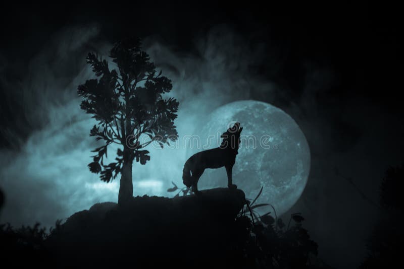 Silhouette of howling wolf against dark toned foggy background and full moon or Wolf in silhouette howling to the full moon. Halloween horror concept. Selective focus. Silhouette of howling wolf against dark toned foggy background and full moon or Wolf in silhouette howling to the full moon. Halloween horror concept. Selective focus