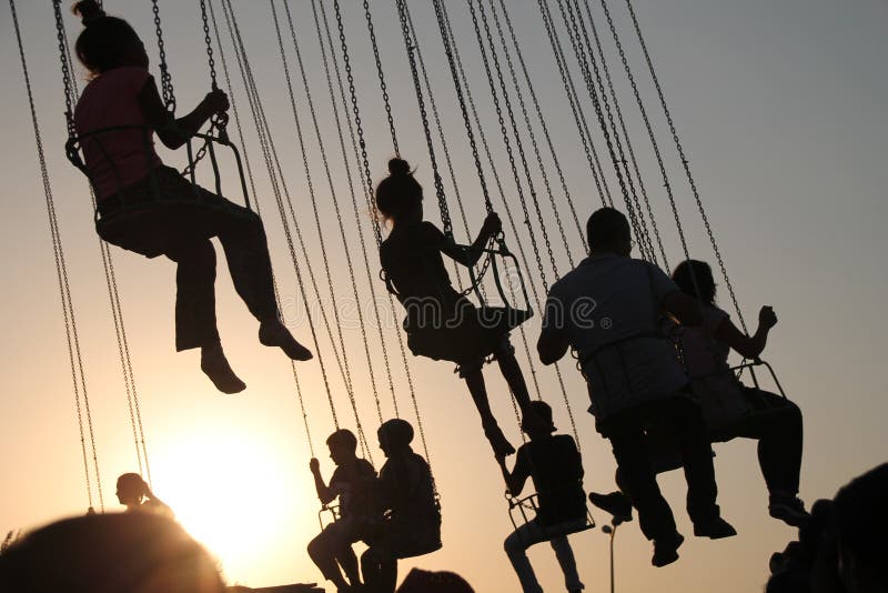 Silueta de la gente joven en la noria y el carrusel de balanceo en el movimiento de parada en fondo de la puesta del sol foto de archivo Silueta de la gente joven en la noria y el carrusel de balanceo en el movimiento de parada en fondo de la puesta del sol foto de archivo