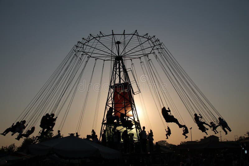 Silueta de la gente joven en la noria y el carrusel de balanceo en el movimiento de parada en fondo de la puesta del sol fotos de archivo Silueta de la gente joven en la noria y el carrusel de balanceo en el movimiento de parada en fondo de la puesta del sol fotos de archivo
