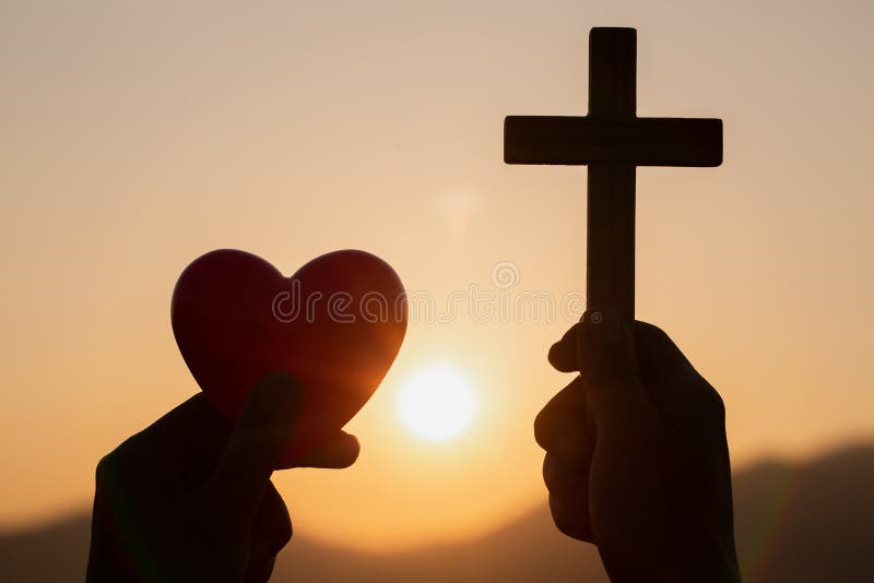 Silueta de las manos de la mujer que ruegan con la cruz y que sostienen una bola roja del corazón en el fondo de la salida del so fotografía de archivo Silueta de las manos de la mujer que ruegan con la cruz y que sostienen una bola roja del corazón en el fondo de la salida del so fotografía de archivo
