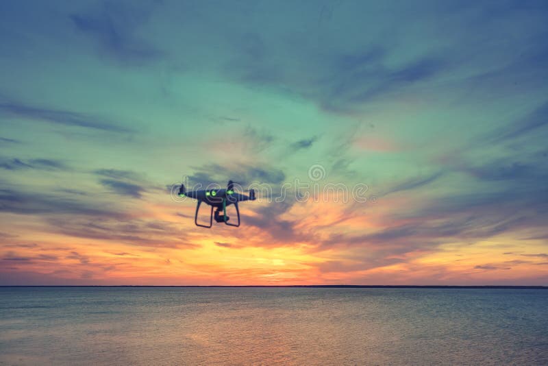 Silhouette of Drone Quad copter flying in sky. Aerial panoramic view of sunset over ocean. Dramatic picturesque evening scene. Colorful cloudy sky in the background. Vehicle at sundown and copy space. Silhouette of Drone Quad copter flying in sky. Aerial panoramic view of sunset over ocean. Dramatic picturesque evening scene. Colorful cloudy sky in the background. Vehicle at sundown and copy space