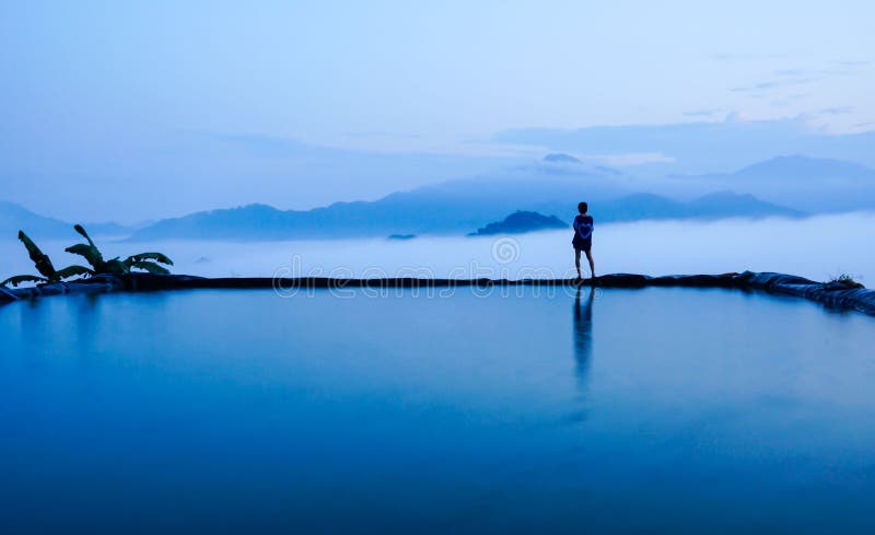 Siluetee la vista posterior de la mujer joven que se coloca cerca de la piscina para el paisaje asombroso del cielo azul y de mon fotos de archivo libres de regalías Siluetee la vista posterior de la mujer joven que se coloca cerca de la piscina para el paisaje asombroso del cielo azul y de mon fotos de archivo libres de regalías