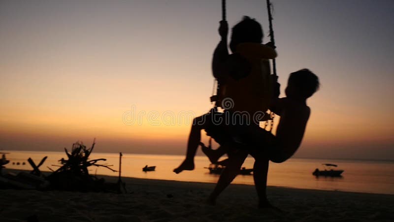 Siluetta di giovane madre che oscilla i piccoli bambini nell'oscillazione della spiaggia alla spiaggia di tramonto Movimento lent