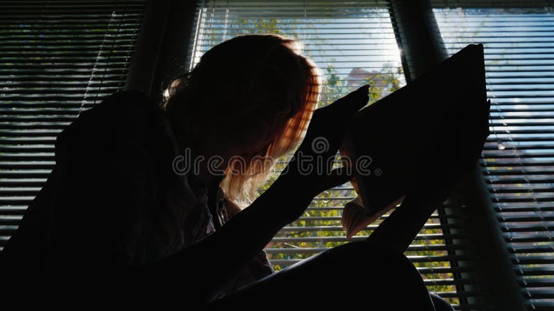 Siluetta di una donna che legge dalla finestra Il sole splende attraverso i ciechi, il chiaro giorno di molla
