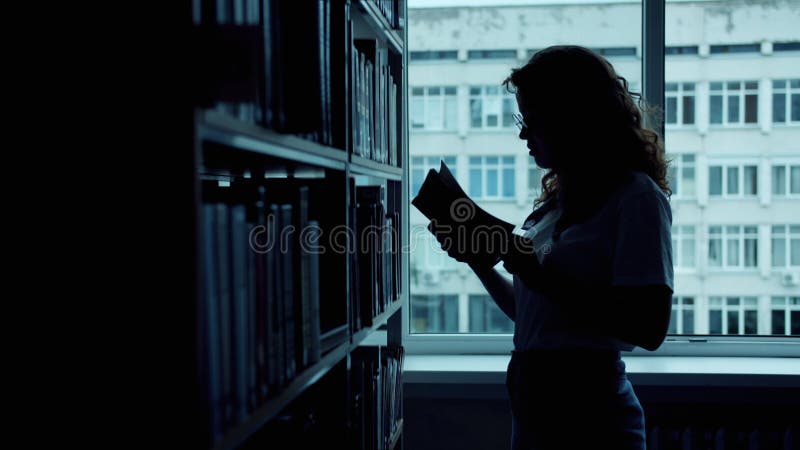 Siluetta di una donna intelligente che sceglie un libro in biblioteca leggendo di mano