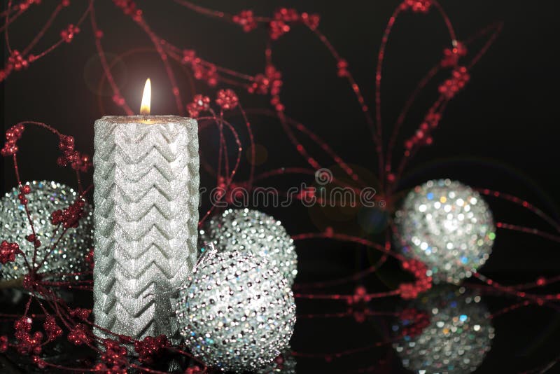 A burning decorative silver candle surrounded by sparkling silver ornaments and sparkling red berry sprays blurred on black background. A burning decorative silver candle surrounded by sparkling silver ornaments and sparkling red berry sprays blurred on black background