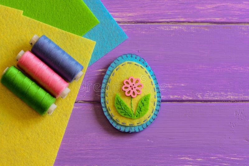 Small felt Easter egg decor with a pink flower, colored thread set, flat felt sheets on a purple wooden background