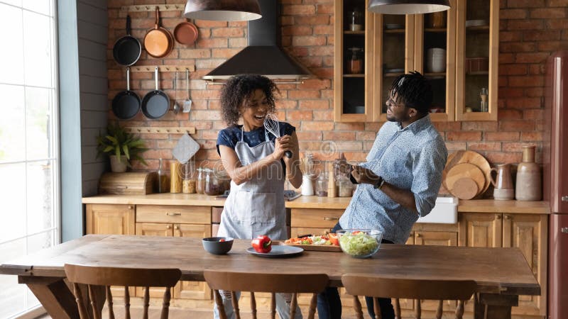 Sing along to my song dear. Joyful millennial black married couple having fun at kitchen sing aloud play music instruments made of kitchenware. Active young black spouses fond of cooking food together. Sing along to my song dear. Joyful millennial black married couple having fun at kitchen sing aloud play music instruments made of kitchenware. Active young black spouses fond of cooking food together