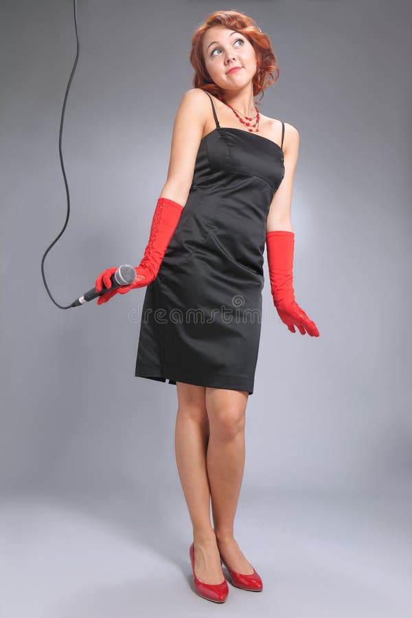 Young woman with microphone standing on grey background in black dress with red accessories. Young woman with microphone standing on grey background in black dress with red accessories.