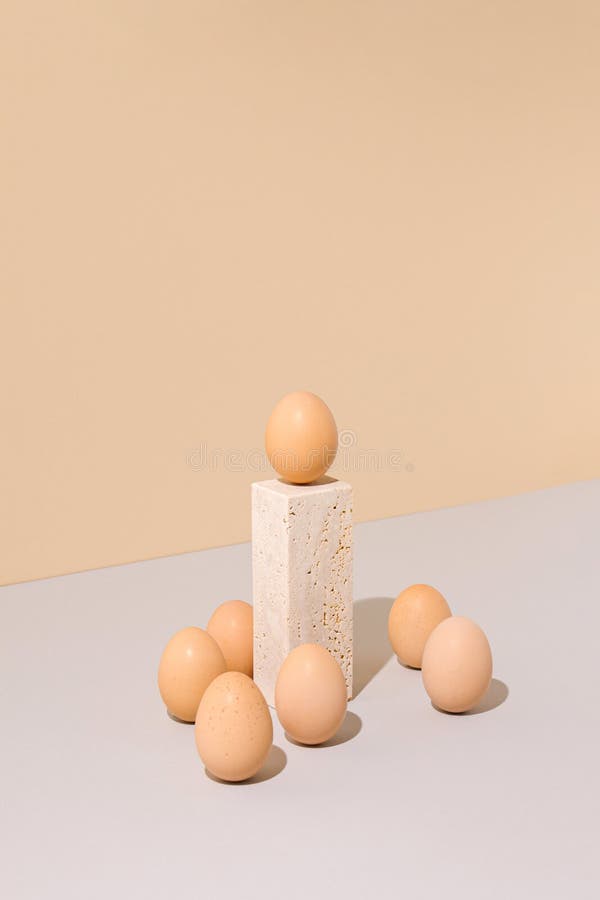 Single egg standing on a travertine marble block podium surrounded by group of eggs on a beige and gray background. royalty free stock image