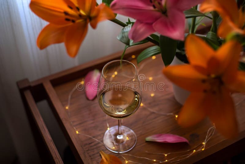 Single glass of white wine, a bouquet of lily flowers and festive lights on a wooden table table. Still life romantic evening stock photo Single glass of white wine, a bouquet of lily flowers and festive lights on a wooden table table. Still life romantic evening stock photo