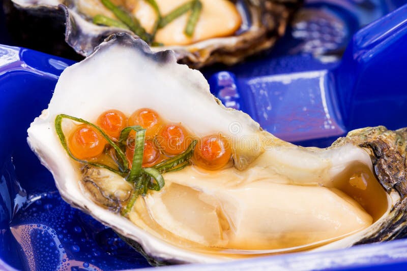 A single oyster live with garnish with an oyster shuck in the foreground with salmon roe