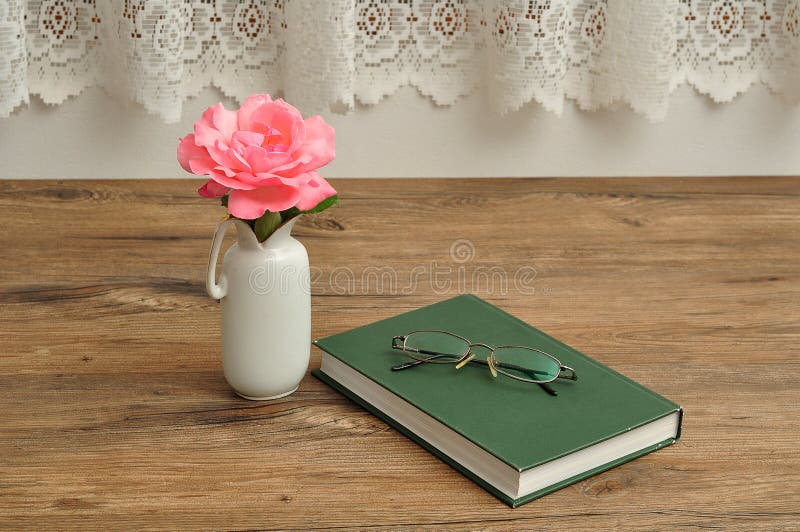 A single pink rose in a vase with a reading book and spectacles