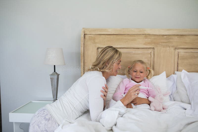 Sira de mãe a verificar a temperatura doente da filha com o termômetro digital no quarto imagens de stock Sira de mãe a verificar a temperatura doente da filha com o termômetro digital no quarto imagens de stock