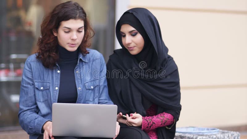 Sitta för två attraktivt unga kvinnor som är utvändigt, och använda bärbara datorn Kors-kulturellt kamratskap Ung muslimkvinna i