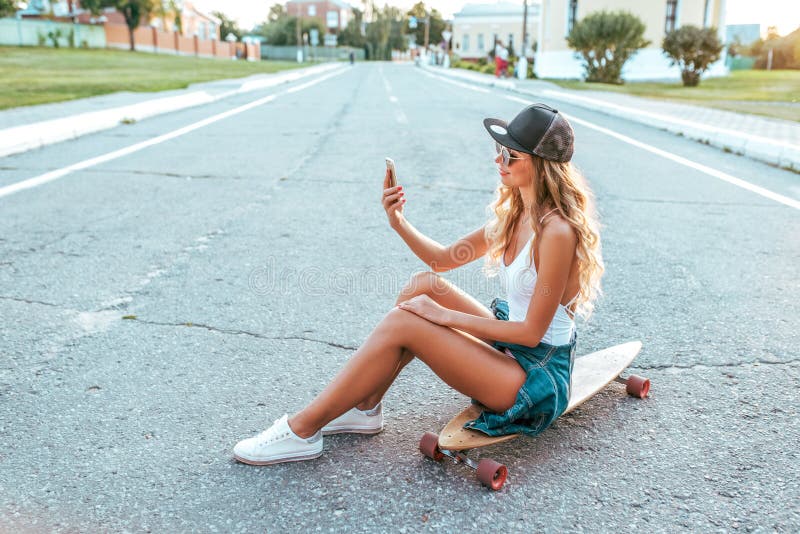 Sittande skateboard för härlig lycklig flicka, fritt utrymme för text Honom som rymmer smartphonen hans hand, talande video appel royaltyfri bild Sittande skateboard för härlig lycklig flicka, fritt utrymme för text Honom som rymmer smartphonen hans hand, talande video appel royaltyfri bild