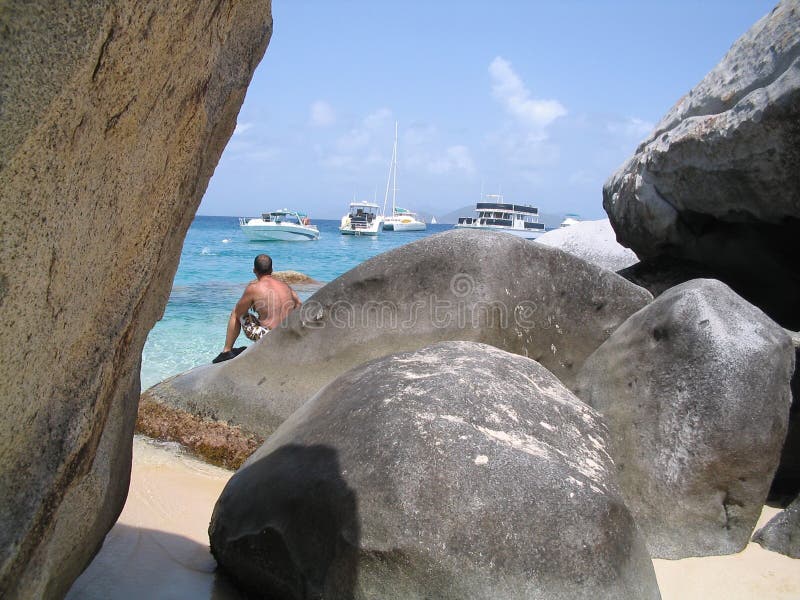 Un uomo seduto su un masso a guardare le barche a vela Devils Bay a Virgin Gorda, Isole Vergini Britanniche.