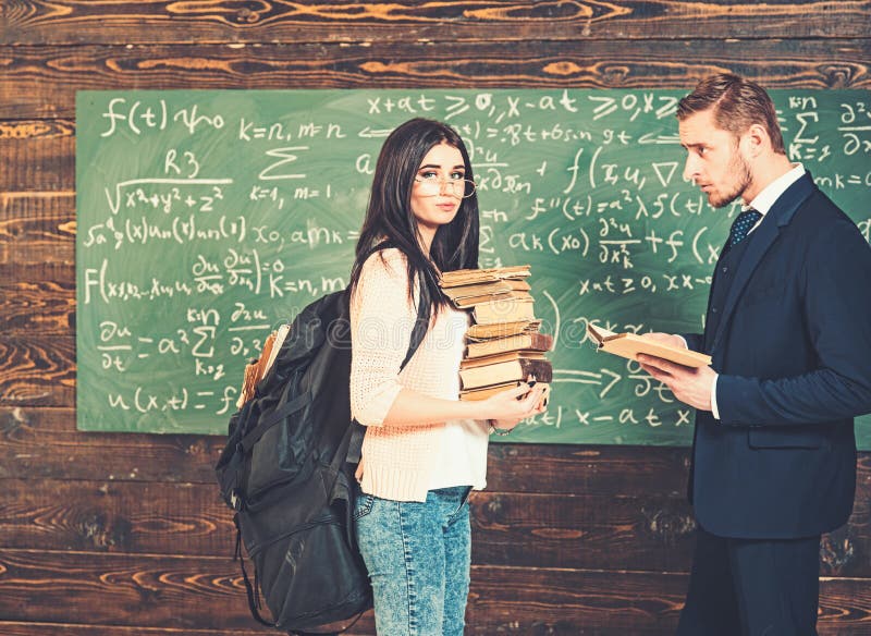 Situaci?n morena del estudiante delante del tablero verde con el mont?n de libros y de la mochila enorme Profesor que mira imagen de archivo libre de regalías Situaci?n morena del estudiante delante del tablero verde con el mont?n de libros y de la mochila enorme Profesor que mira imagen de archivo libre de regalías