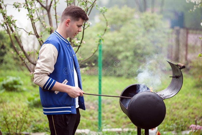 Situación del hombre joven cerca de la parrilla de la barbacoa Inflamaci?n del fuego en el aire abierto comida campestre del vera imagen de archivo libre de regalías Situación del hombre joven cerca de la parrilla de la barbacoa Inflamaci?n del fuego en el aire abierto comida campestre del vera imagen de archivo libre de regalías